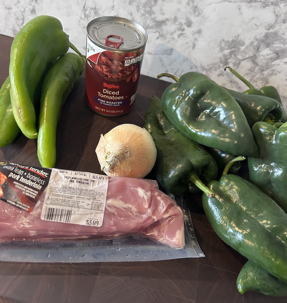 Anaheim peppers, poblano peppers, an onion, a can of fire roasted tomatoes and a pork tenderloin on a wooden cutting board for making smoked green chili.