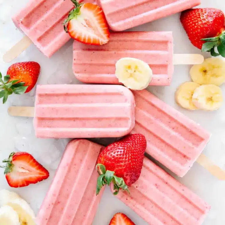 A bunch of sugar free strawberry banana popsicles on a marble background with fresh banana slices and halved strawberries.
