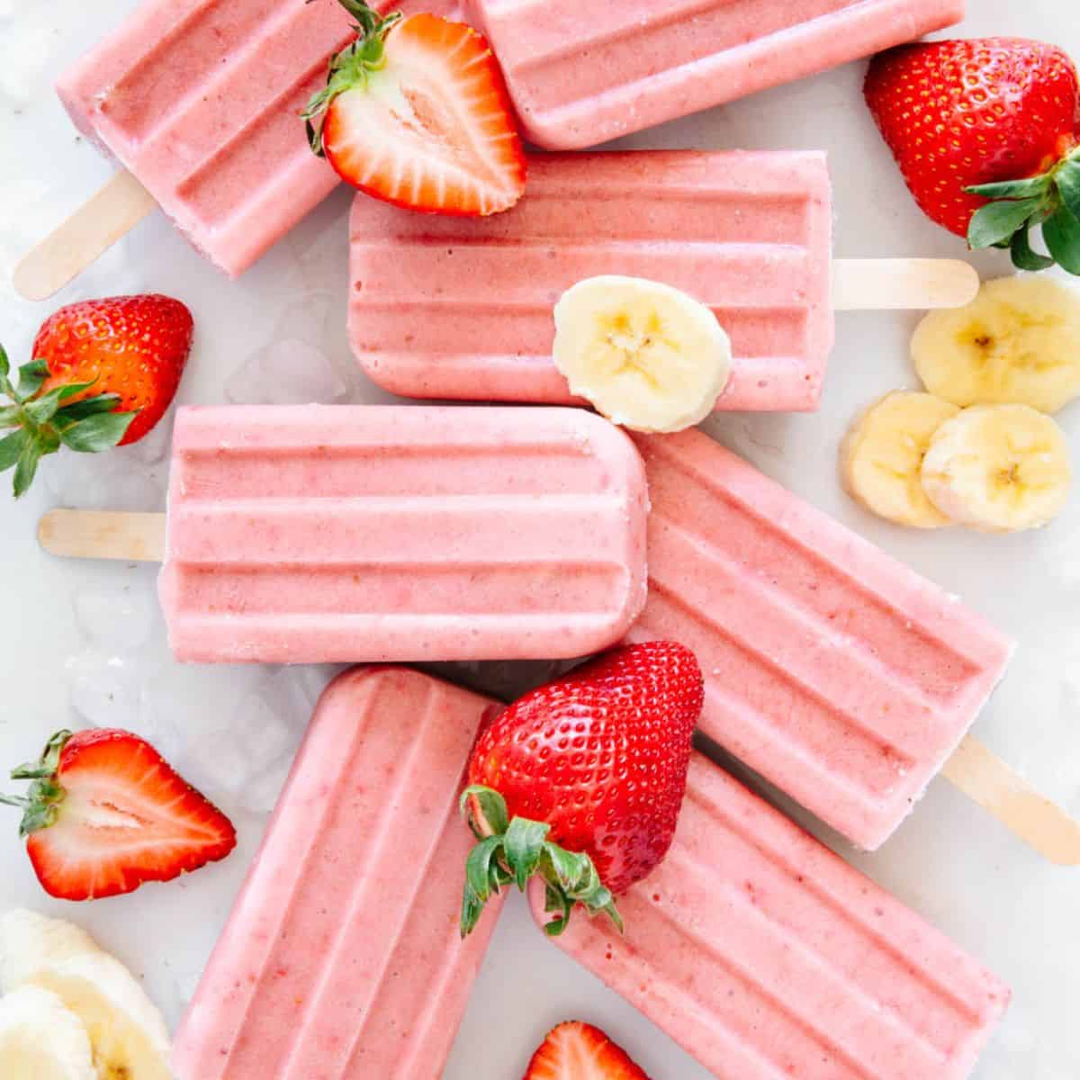A bunch of sugar free strawberry banana popsicles on a marble background with fresh banana slices and halved strawberries.