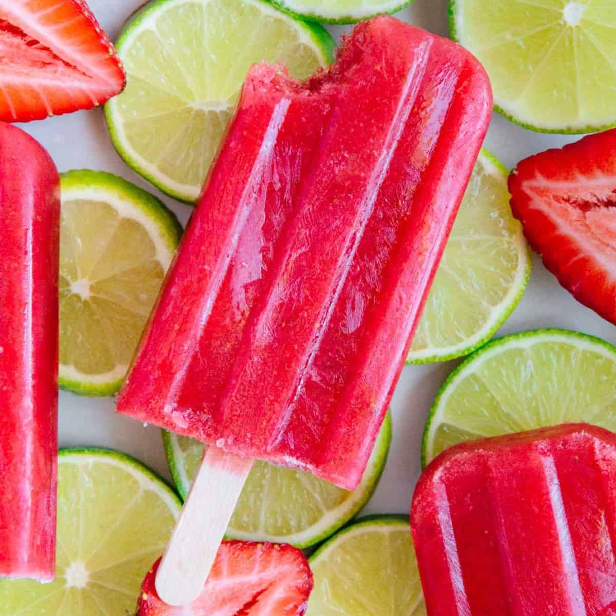 A strawberry lime popsicle with a bite out of it resting on top of fresh lime slices and fresh strawberry halves.