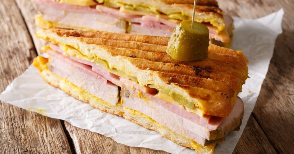 A cuban sandwich on a paper wrapper on a wooden table with a pickle.