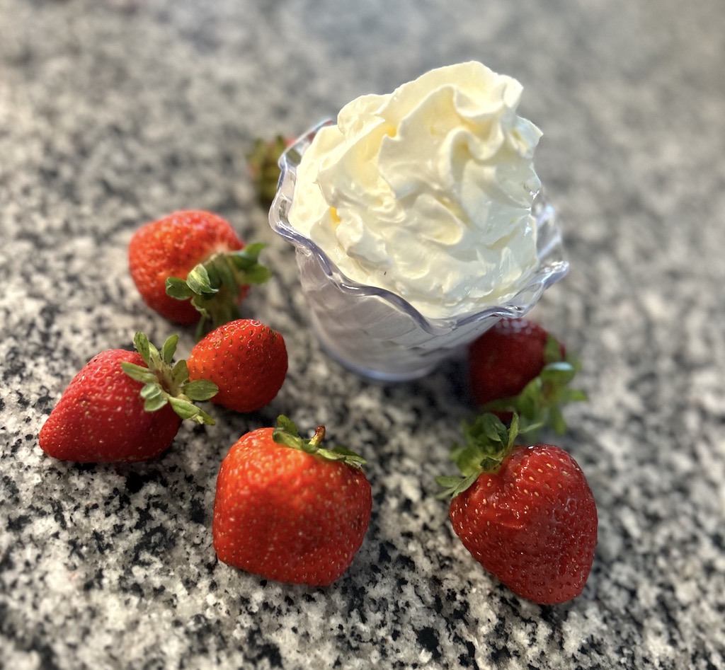 A small clear bowl of whipped cream on a marble counter top with fresh strawberries.