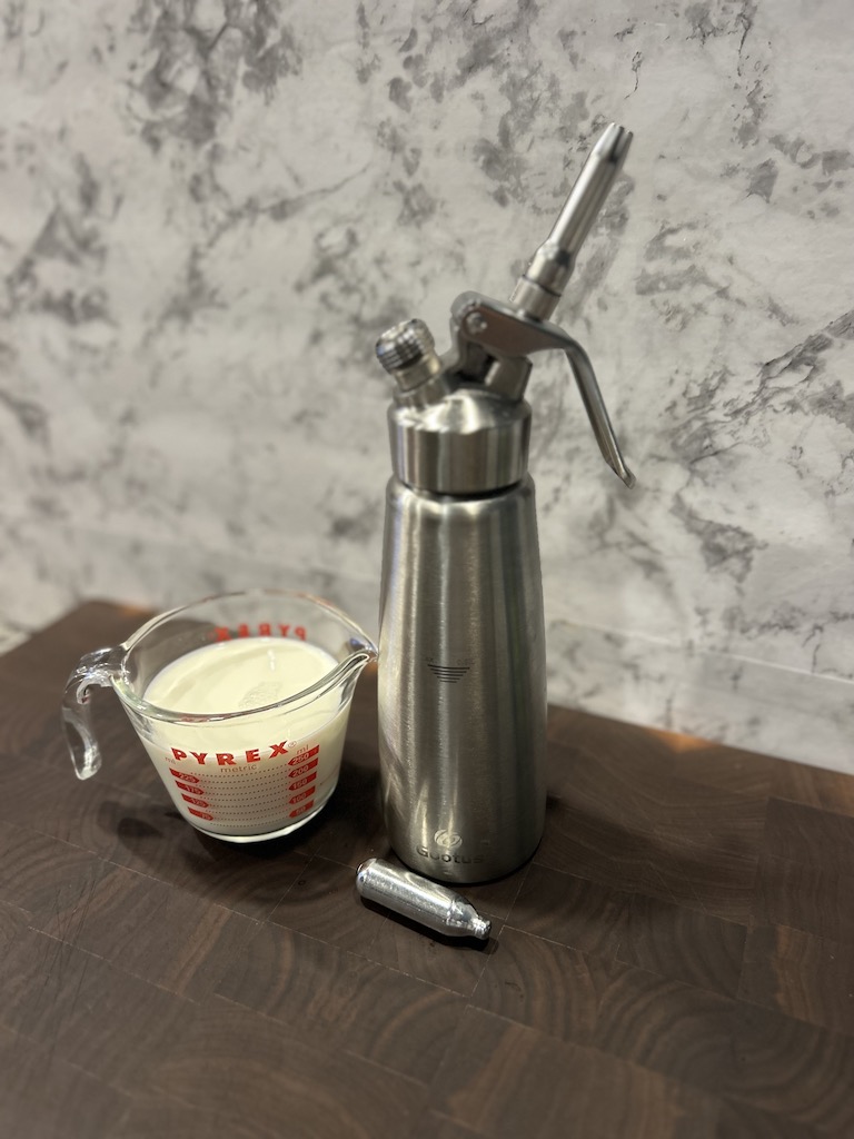 A whipped cream dispenser, a N20 cartridge, and a cup of heavy cream on a wooden cutting board with a marble background for making whipped cream dispenser recipes.