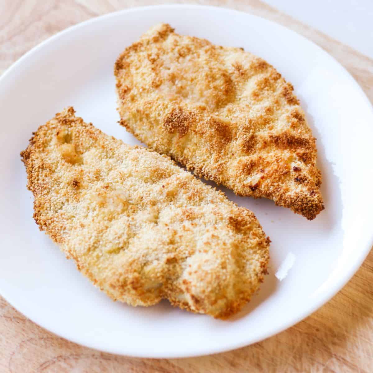 Two air fryer breaded chicken cutlets on a white plate on a wooden counter.