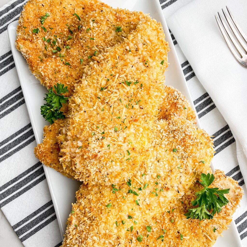 13 Recipes for Thinly Sliced Chicken Breasts Thin chicken breasts coated with a crispy breading baked and plated on a long rectangle white plate on a stripped napkin.