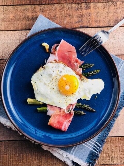 A blue plate featuring eggs and asparagus with a fork on a wooden table.
