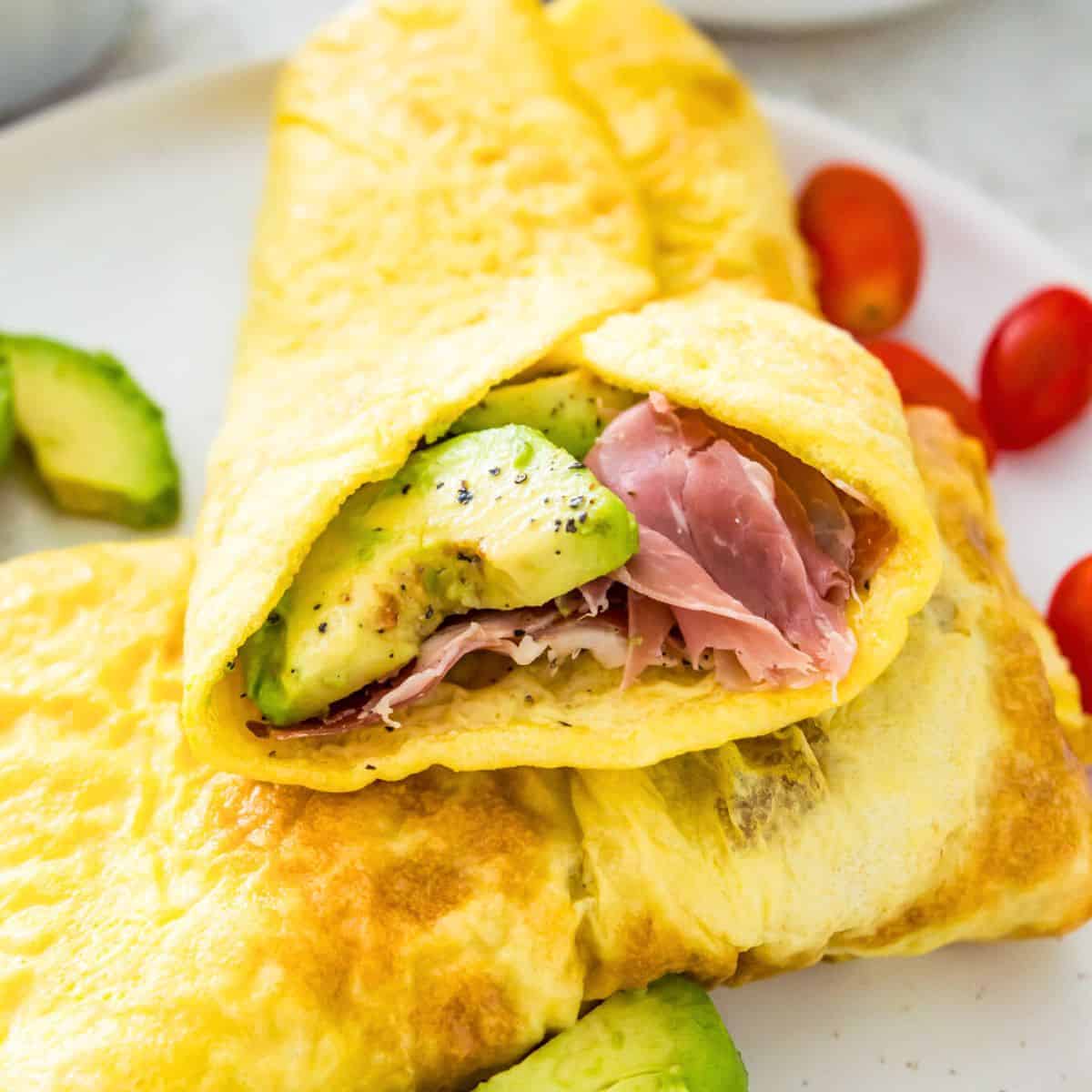 A delicious egg wrap filled with avocado and ham on a white plate with fresh avocado slices and cherry tomatoes.