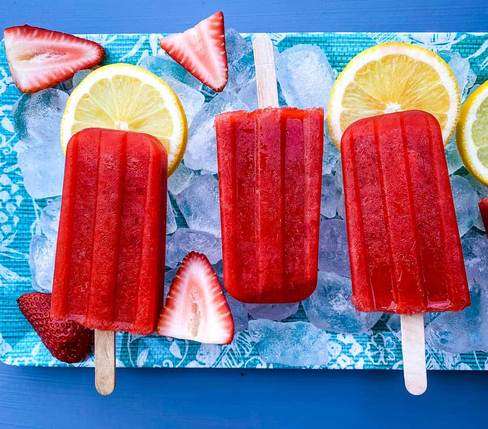 Strawberry lemon popsicles on ice on a blue background with fresh lemon slices and halved strawberries.
