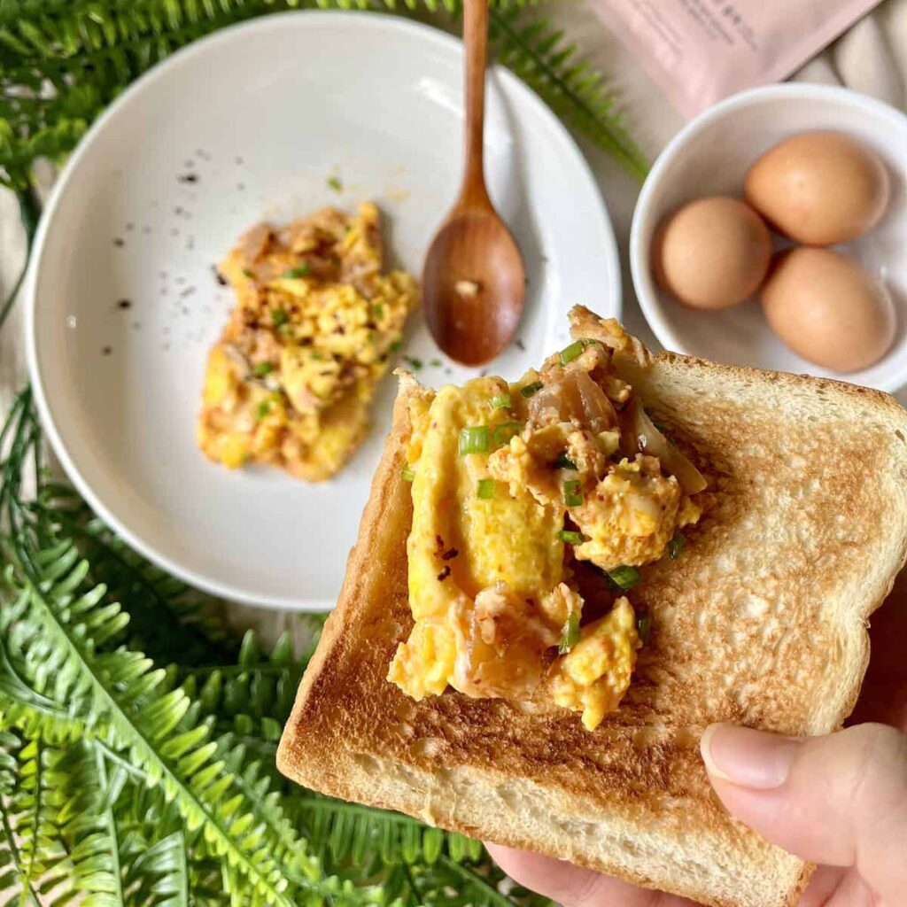 A woman's hand holding a piece of kimchi egg toast over a bowl of kimchi eggs.