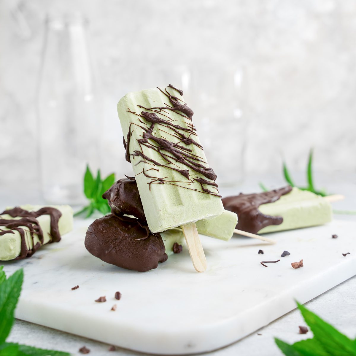 A sugar free matcha mint chocolate popsicle resting o top of a few other mint chocolate chip popiscles on a marble background with fresh mint leaves.