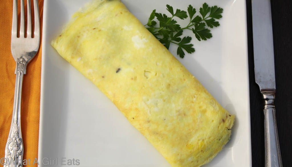 The perfect french omelet on a white plate garnished with fresh parsley with a fork and knife on either side.