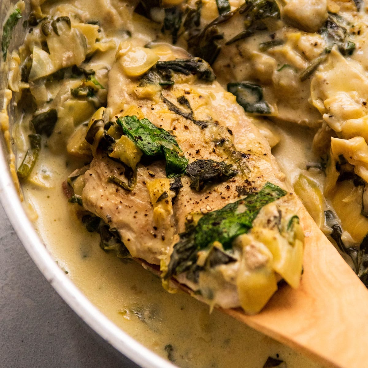 A white bowl filled with spinach artichoke chicken with a wooden spoon dishing up a serving.