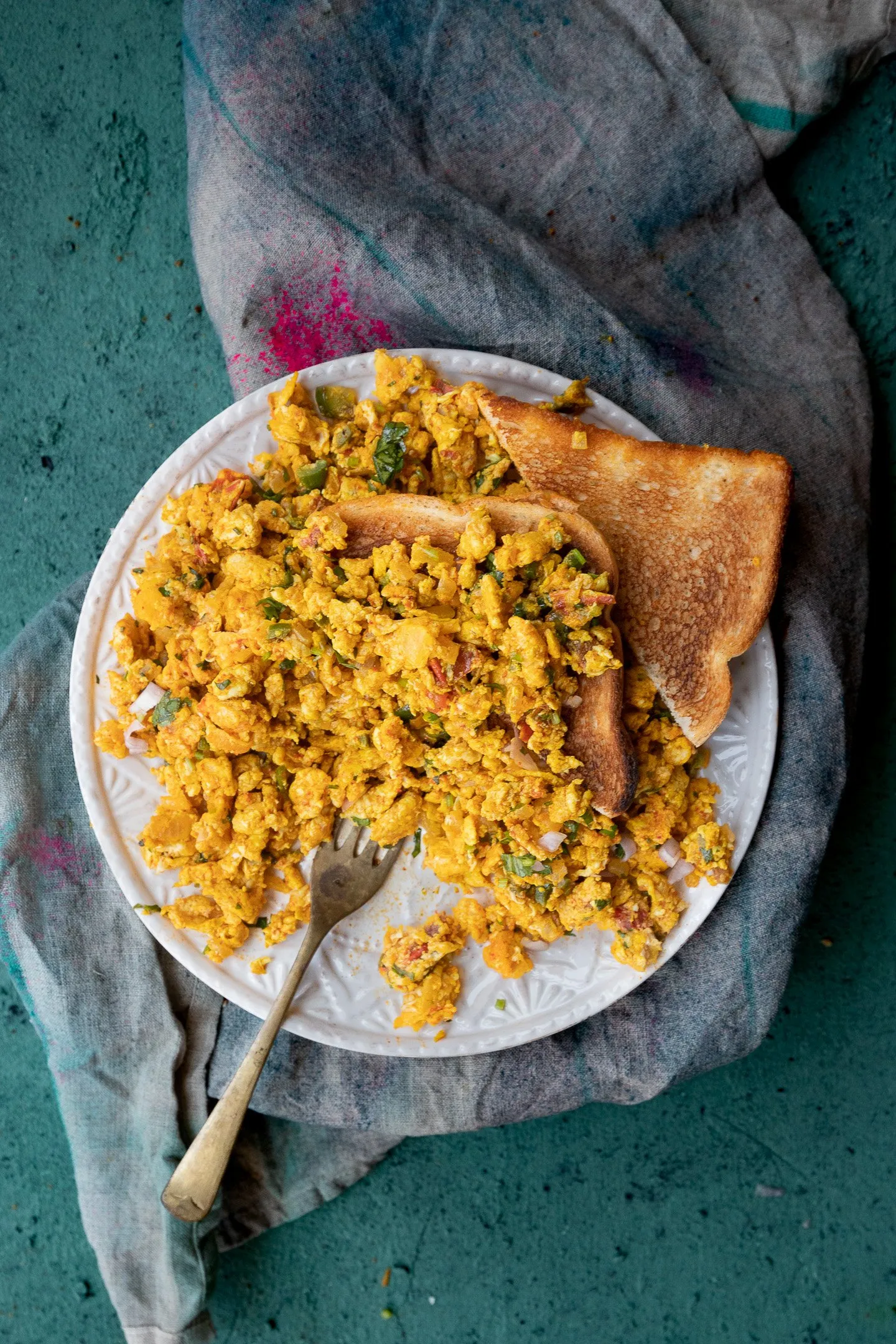 Egg bhurji on a white plate with a fork and two pieces of toast on a cloth napkin.