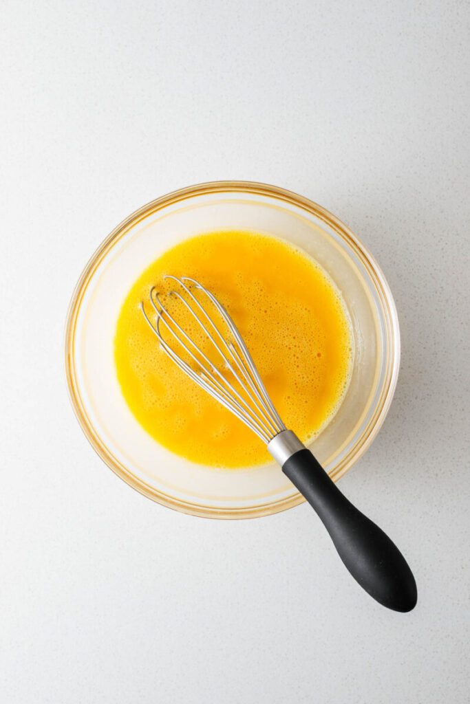 A glass bowl filled with whisked eggs and a whisk.