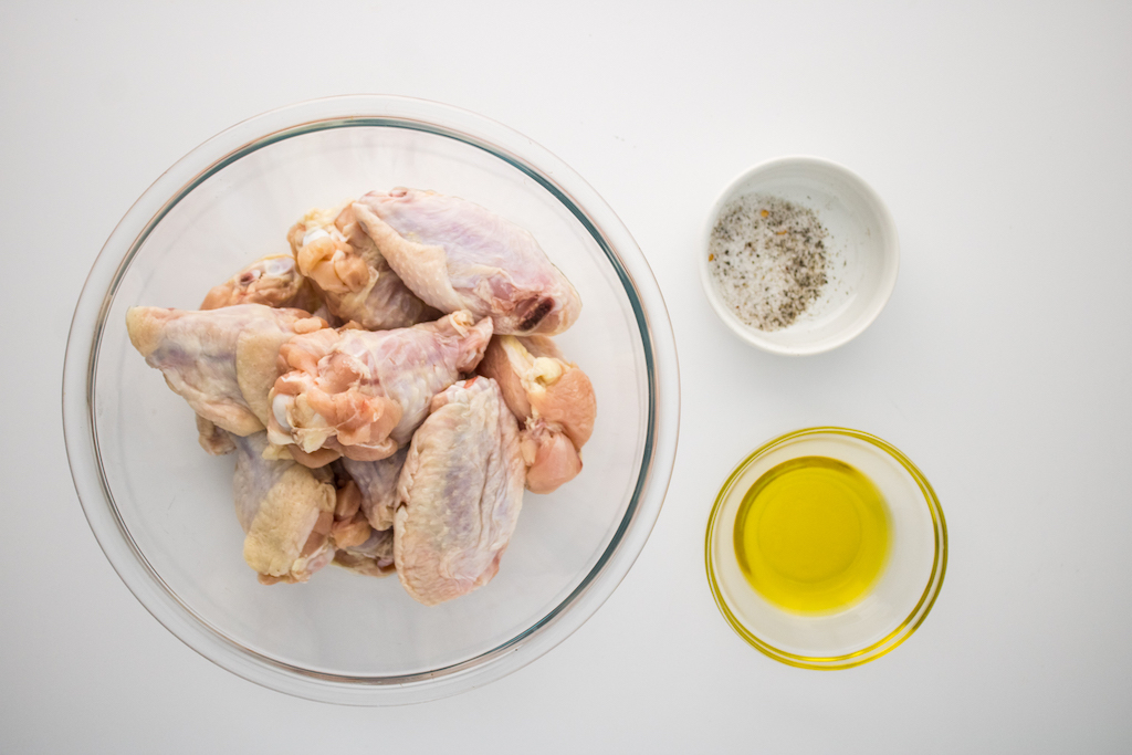A glass mixing bowl filled with chicken wings next to small glass bowls of olive oil and salt and pepper on a white counter.