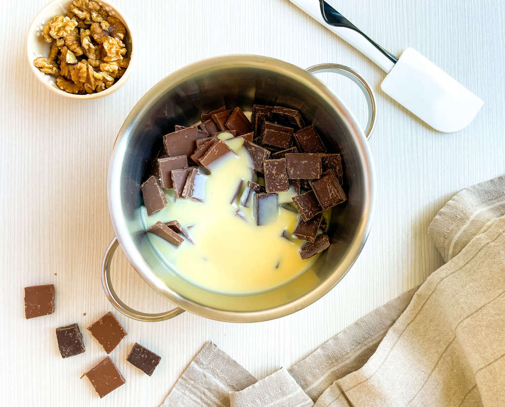 Milk chocolate, dark chocolate, and sweeteneed condensed milk in a saucepan with a cup of walnuts and spatula next to it to make Walnut Chocolate Fudge.