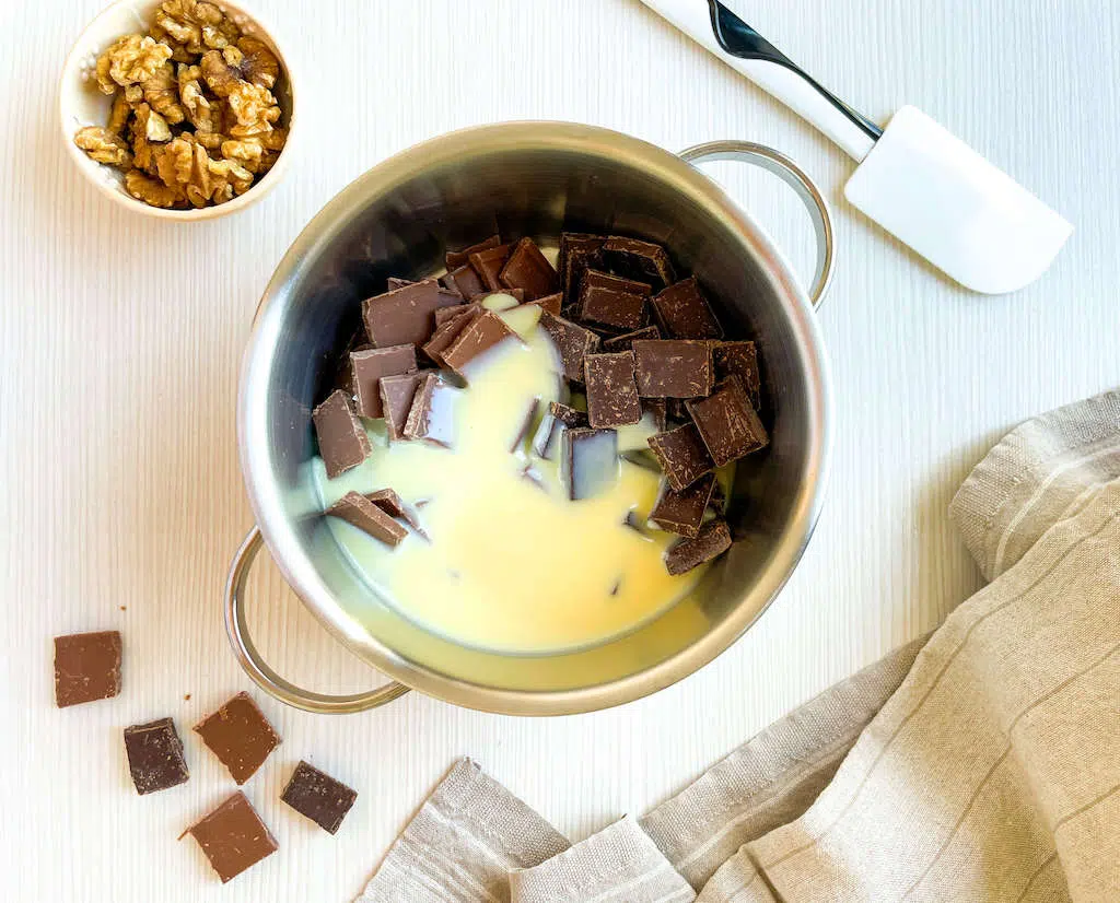 Milk chocolate, dark chocolate, and sweeteneed condensed milk in a saucepan with a cup of walnuts and spatula next to it to make Walnut Chocolate Fudge.