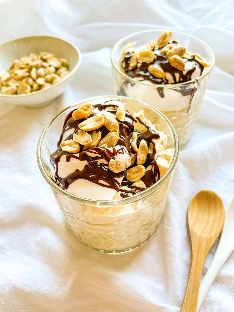 Two parfait glasses of chocolate peanut butter mousse on a white cloth with a wooden spoon and small bowl of chopped peanuts.