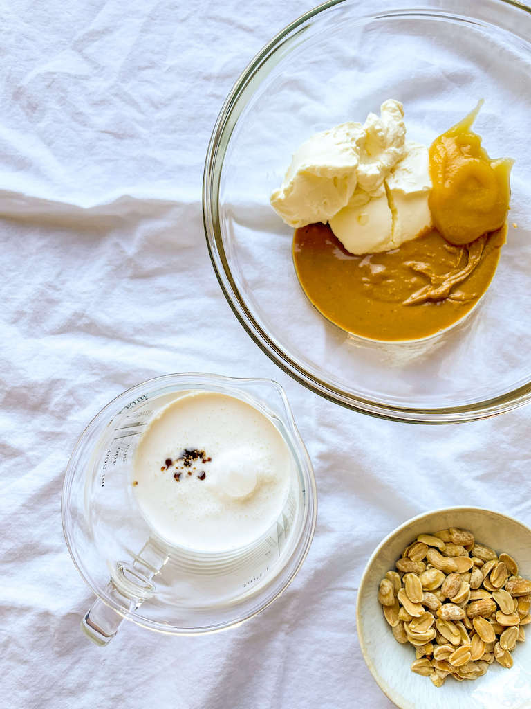 Peanut Butter, honey and cream cheese in a glass mixing bowl on a white cloth with heavy cream and vanilla extract in a measure cup.