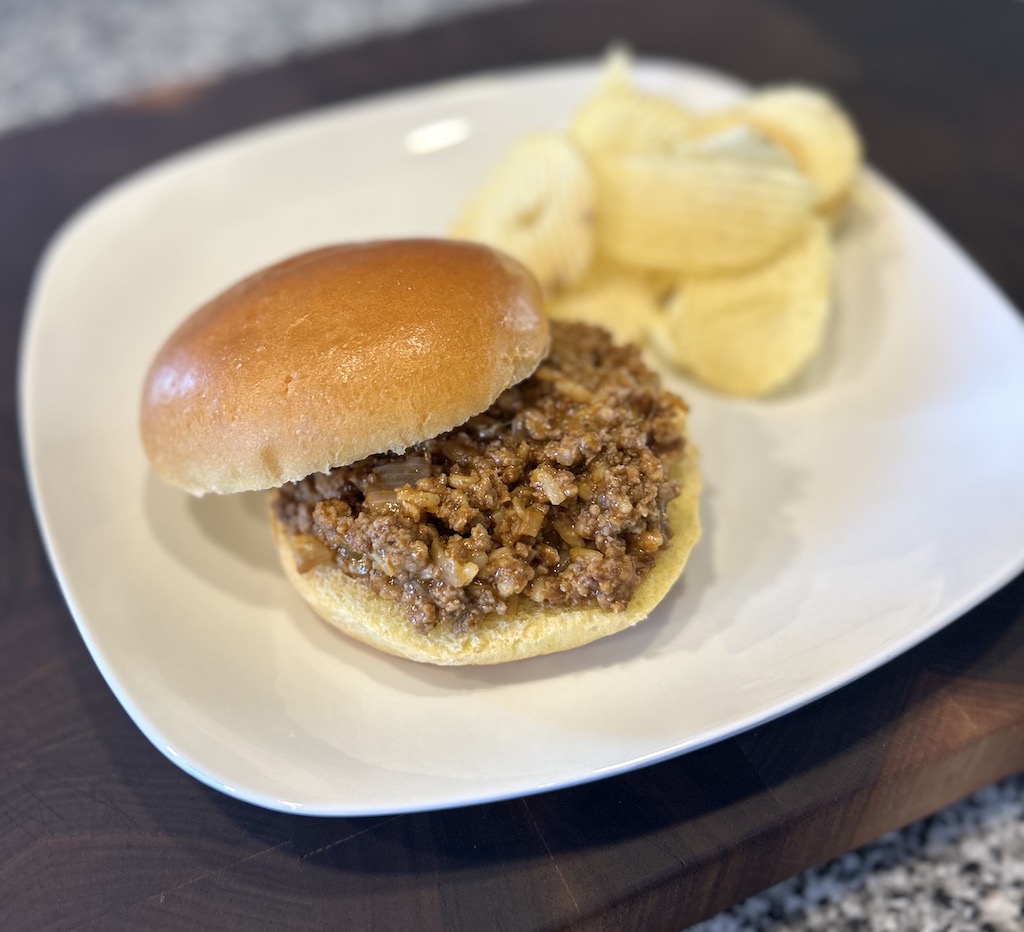 Chicken gumbo sloppy joe on a hamburger bun served on a white plate with chips.