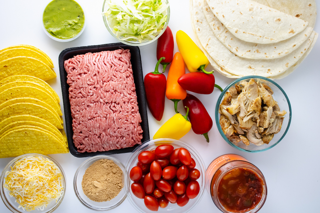 The ingredients for a taco charcuterie board including ground beef, peppers, lettuce, taco shells, tortillas, guacamole and chicken.