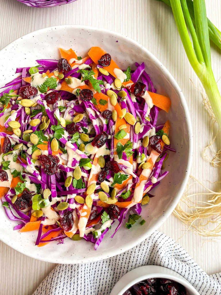 Carrot and red cabbage salad with lemon tahini dressing garnished with pumpkin seeds, dried cranberries and fresh parsley in a stoneware bowl.