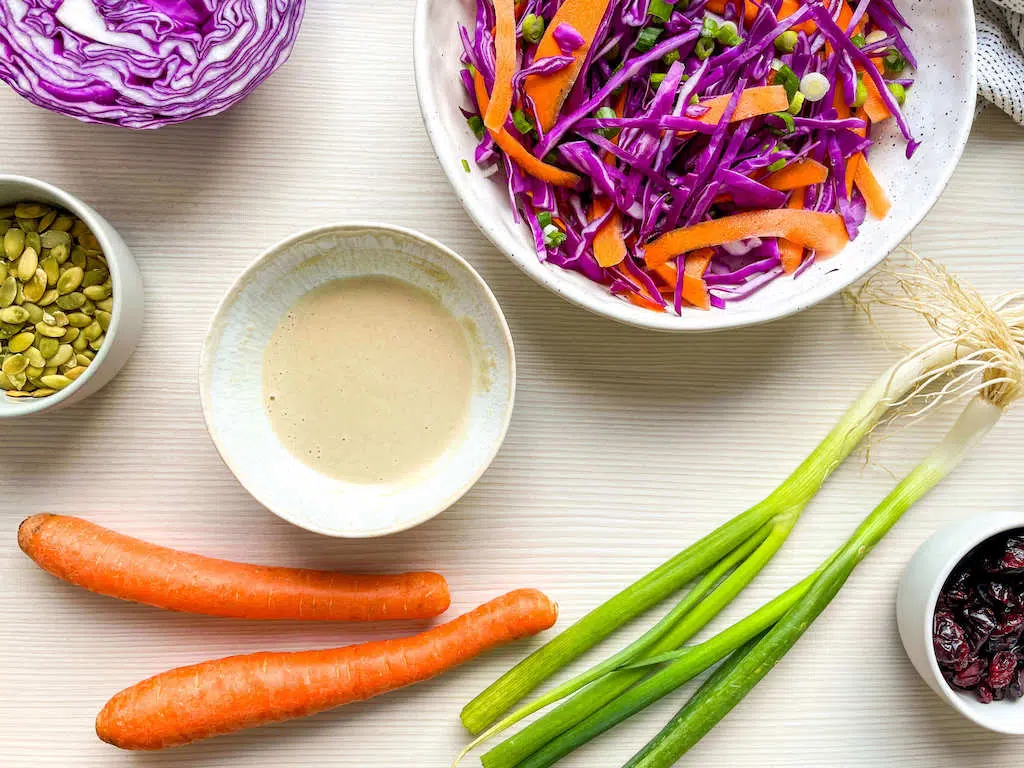 Thinly sliced red cabbage, carrots and green onions tossed together in a stoneware bowl next to a small bowl of lemon tahini dressing.