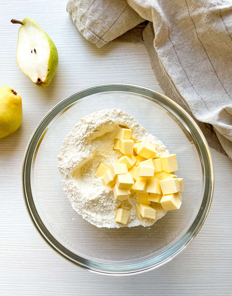 Cold cubed butter in the mixing bowl with the flour next to a half of a pear.