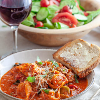 A bowl of Italian Sausage and Tortellini with a side of garlic bread and a garden salad next to a glass of red wine.