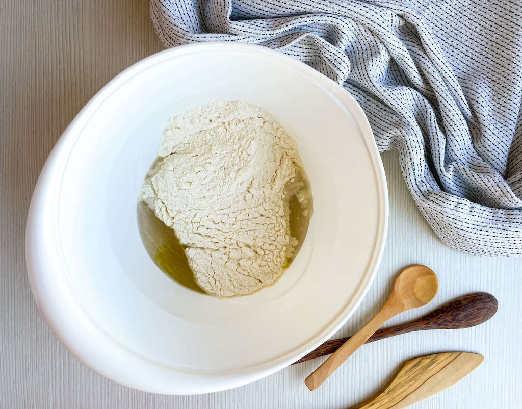 Adding the flour to the yeast mixture in a white mixing bowl.