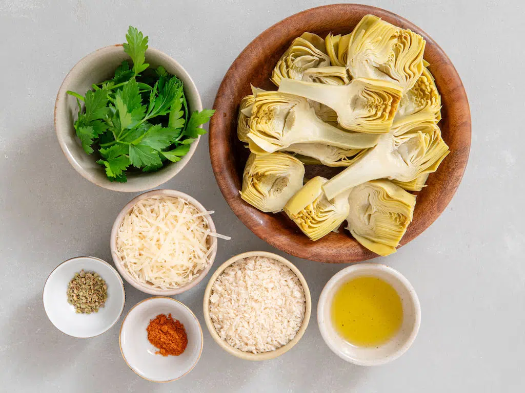 The ingredients for crispy artichoke hearts in small bowls including canned artichoke hearts, parmesan cheese, olive oil, paprika, thyme, panko and fresh parsley.