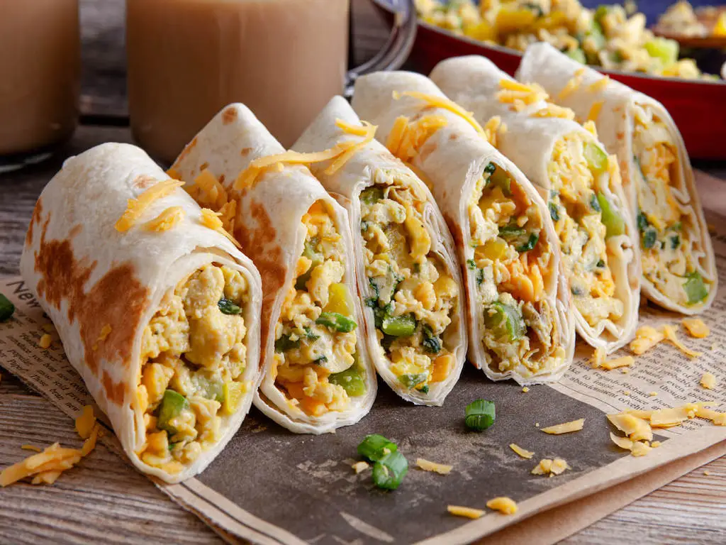 Six breakfast burritos garnished with shredded cheese on a table with a newspaper.