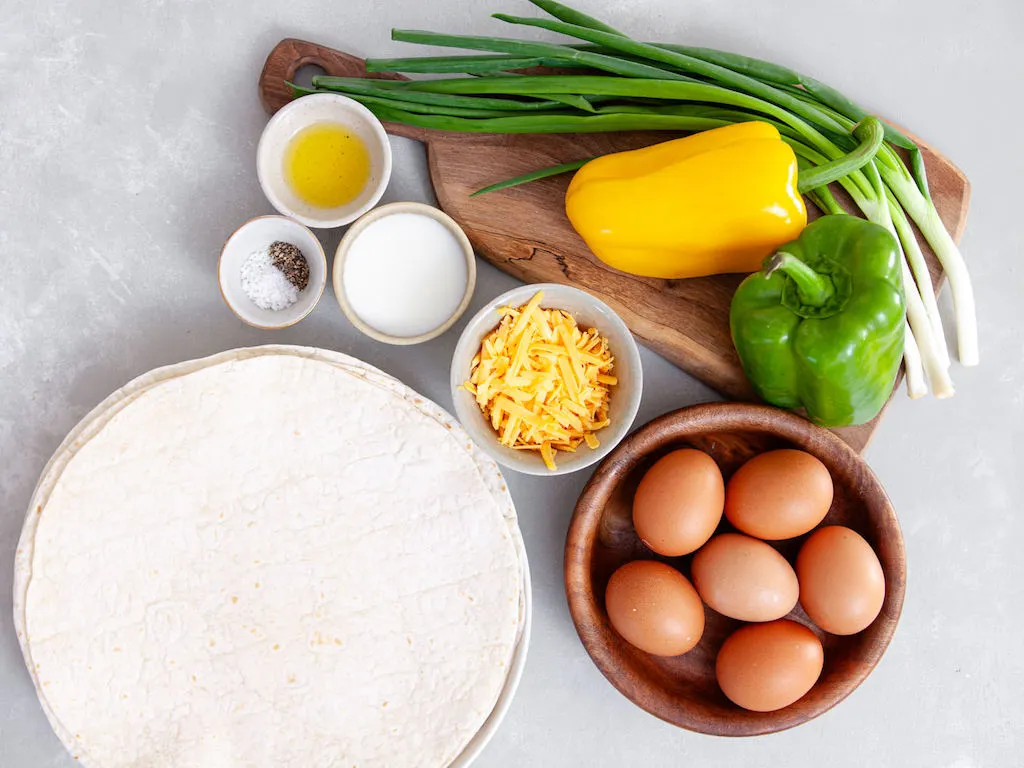 Green onions, bell peppers, shredded cheese, eggs, milk, olive oil, and tortillas artfully displayed on a counter.