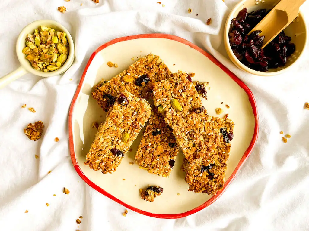 A plate of pistachio granola bars on a white napkin with a bowl of dried cranberries and pistachios.