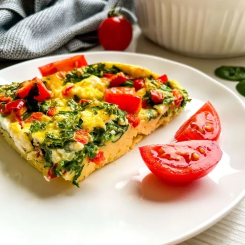 A slice of spinach feta breakfast casserole on a plate garnished with sliced tomatoes.