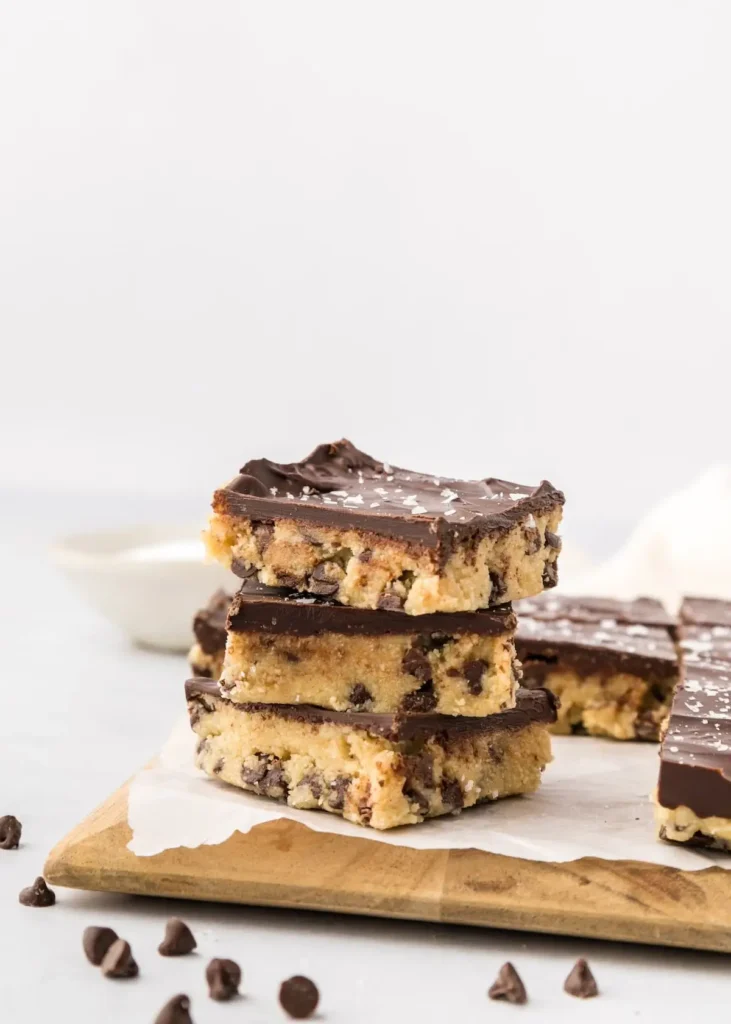 Stacked cookie dough bars loaded with chocolate chips, featuring a rich chocolate topping and a sprinkle of sea salt.