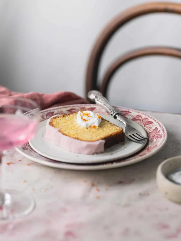  A light and tender olive oil cake topped with a pink orange glaze, served with a dollop of whipped cream on a vintage plate for a classic yet elegant dessert.