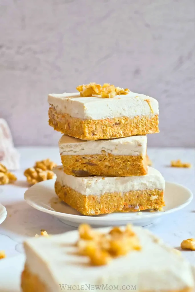   No-bake carrot cake bars topped with a creamy cashew frosting, garnished with chopped walnuts, and layered with wholesome ingredients, perfect for healthy indulgence.