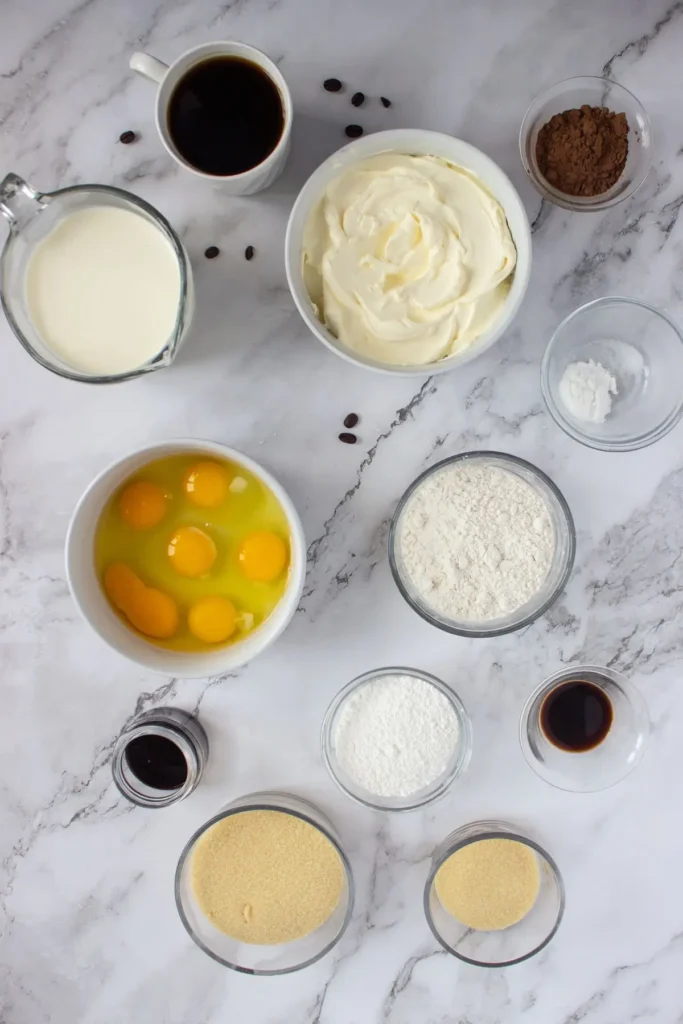 A flat lay of ingredients for an easy tiramisu recipe, including eggs, mascarpone, coffee, cocoa powder, sugar, and flour, arranged on a marble surface.