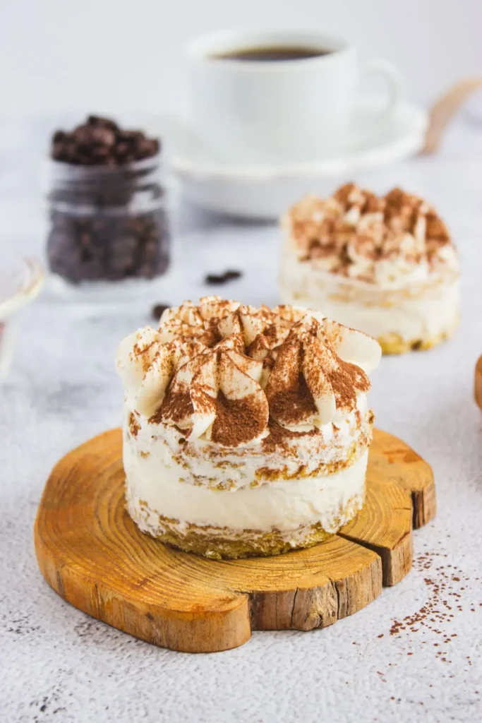 Mini tiramisu desserts served on wooden boards, topped with whipped mascarpone and dusted with cocoa powder, with a jar of coffee beans in the background.