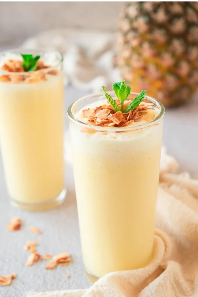 Two glasses of frothy pineapple shake topped with coconut flakes and mint, alongside a fresh pineapple. A healthy and delicious nonalcoholic pineapple drink.