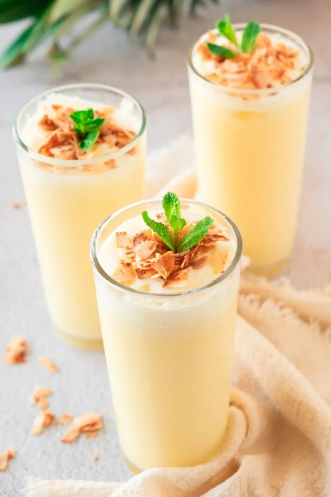 Three glasses of frothy pineapple shake topped with coconut shavings and mint sprigs, alongside a whole pineapple. A delicious and nutritious nonalcoholic pineapple drink for all ages.