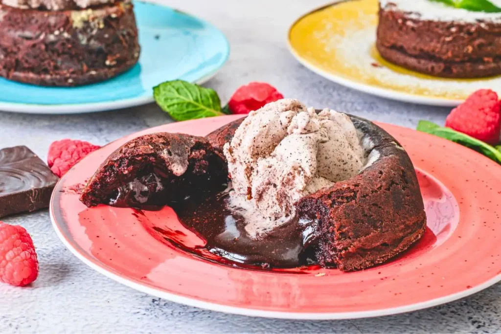 Molten Lava Chocolate Cake Molten lava chocolate cake on a pink plate, cut open with gooey chocolate, topped with ice cream, raspberries, and mint leaves, with other cakes on colorful plates.
