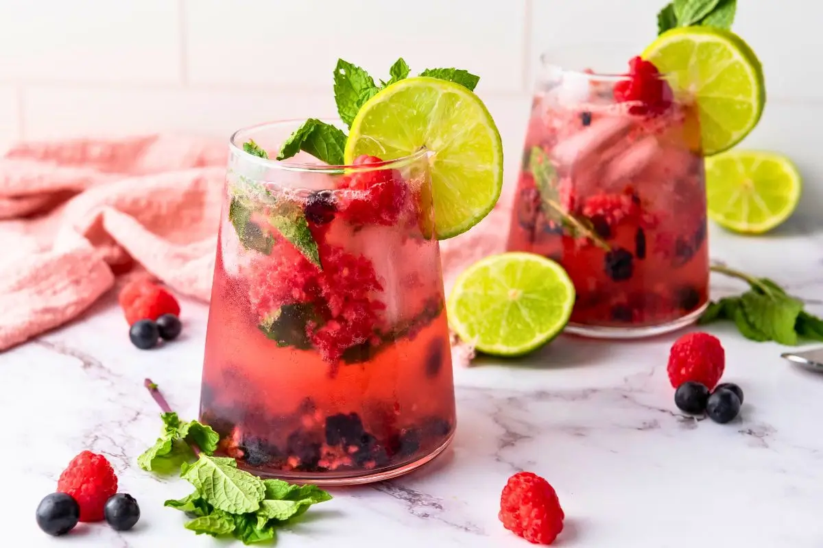 Two glasses of a finished blueberry raspberry mint mocktail with mint sprigs and lime wedges, a fun non alcoholic berry drink ready to serve.