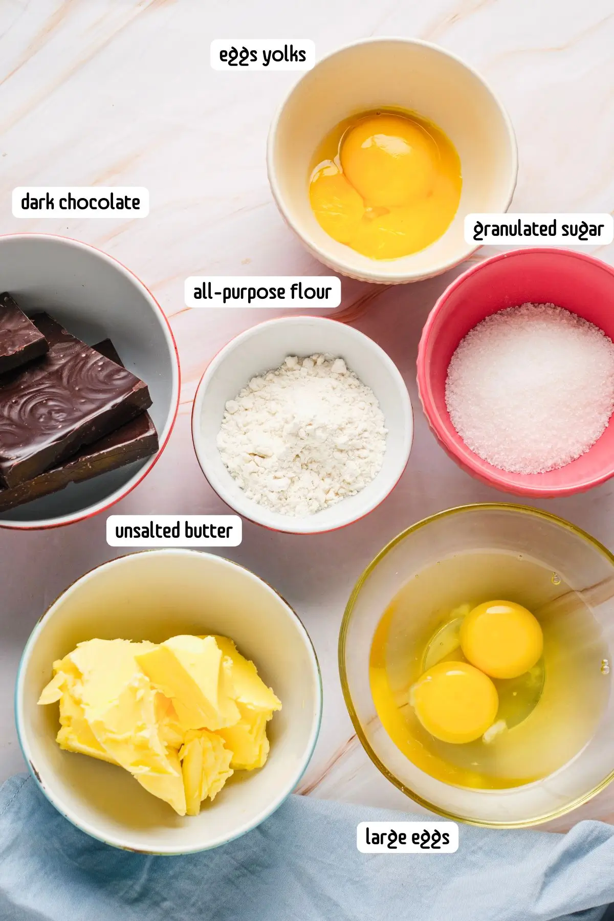 Molten Lava Chocolate Cake Bowls of ingredients including dark chocolate, flour, sugar, eggs, egg yolks, and butter arranged on a marble countertop.