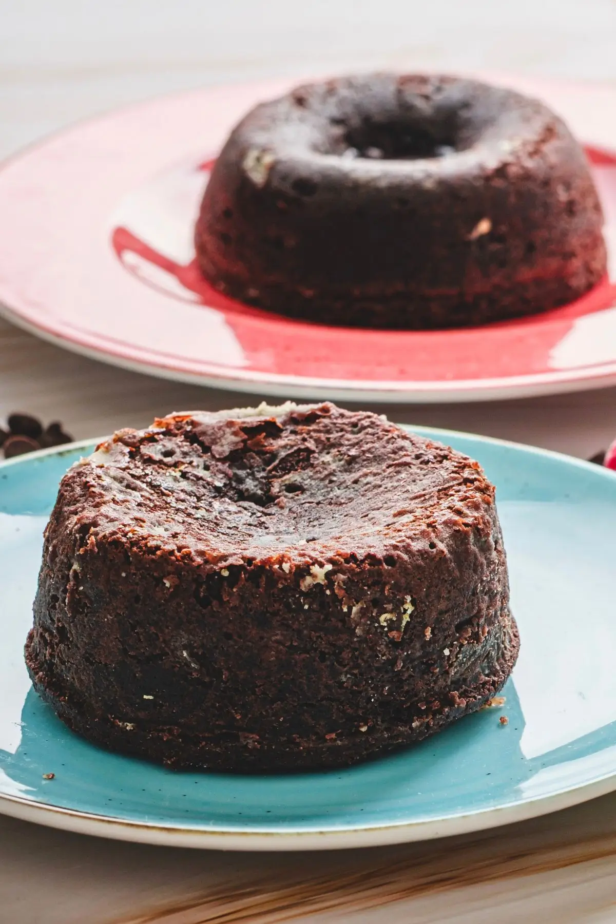 Molten Lava Chocolate Cake Molten lava chocolate cake on a blue plate, uncut with a textured top, next to a similar cake on a pink plate, with chocolate chips on a wooden surface.