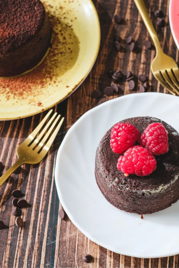 Molten lava chocolate cake on a white plate with three raspberries on top, served with a gold fork on a wooden table.