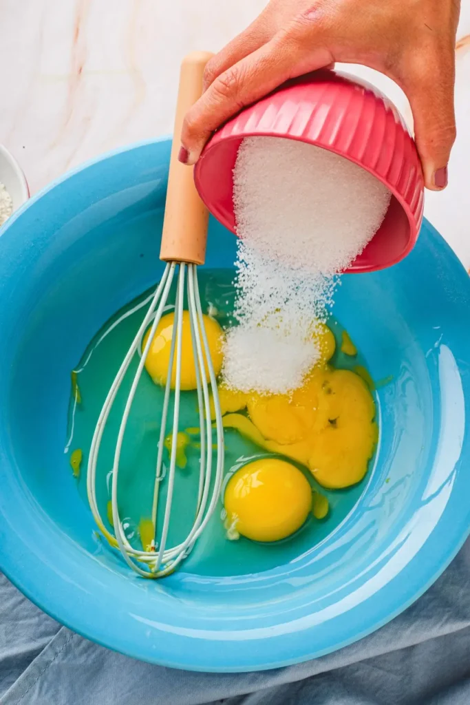 Hand adding sugar to eggs in a blue bowl with a whisk, with bowls of ingredients like butter and chocolate in the background.