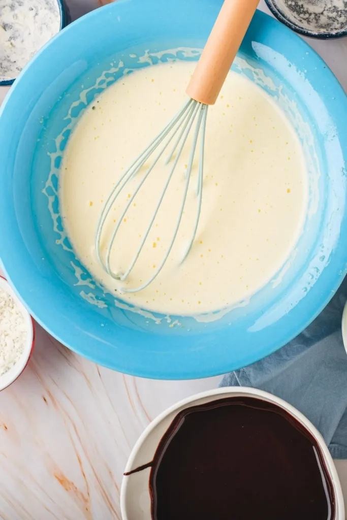 Whisked pale egg mixture in a blue bowl with a whisk, surrounded by bowls of flour and melted chocolate on a marble surface.