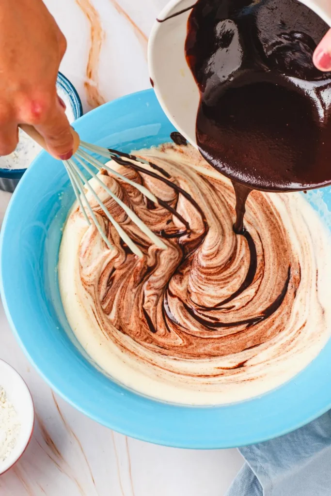 Hand pouring melted chocolate into a blue bowl with whisked egg mixture, creating a swirled texture.